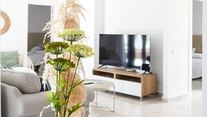 Una sala moderna con un sofá blanco, una mesita pequeña blanca, un televisor sobre un mueble de madera y una planta decorativa con hojas verdes y plumas blancas.
