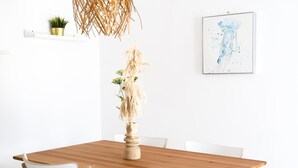 Una mesa de comedor con sillas blancas, un jarrón con plantas secas, una lámpara colgante y un cuadro abstracto enmarcado en la pared.