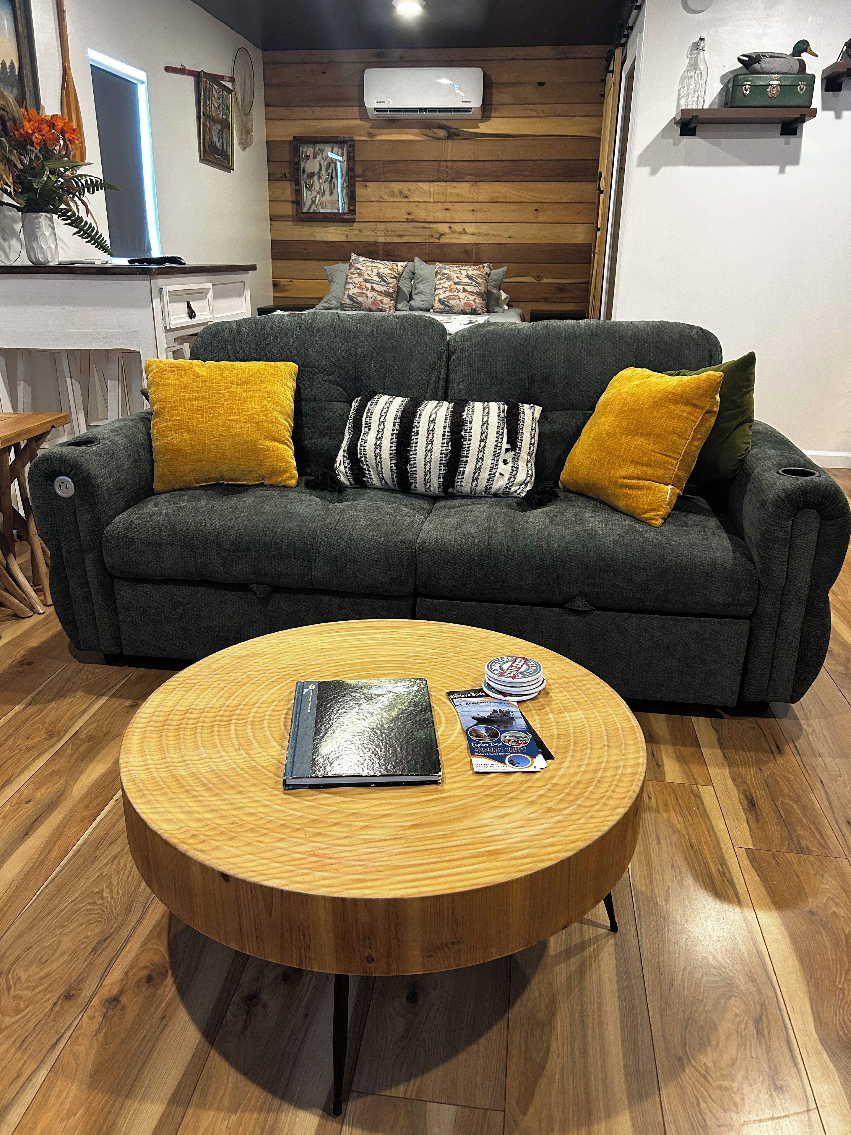 Un salon avec un canapé vert foncé, deux coussins jaunes, une table basse ronde en bois sur laquelle reposent un livre et un magazine, et un plancher en bois.