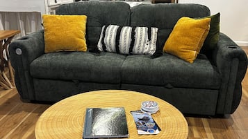 Un salon avec un canapé vert foncé, deux coussins jaunes, une table basse ronde en bois sur laquelle reposent un livre et un magazine, et un plancher en bois.