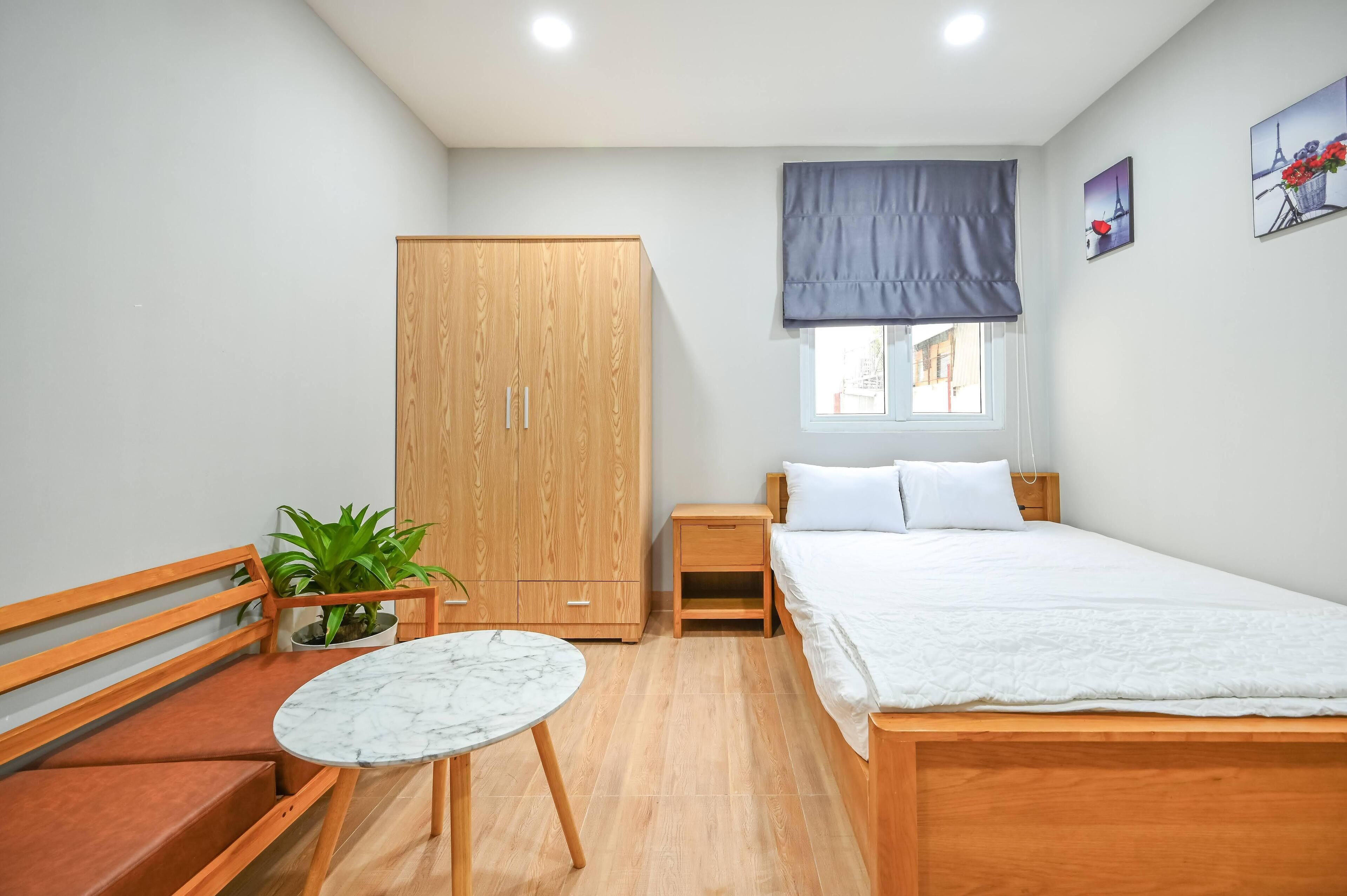 A bedroom with a wooden bed, a nightstand, a wooden wardrobe, a small table with a plant, and a window with a view.