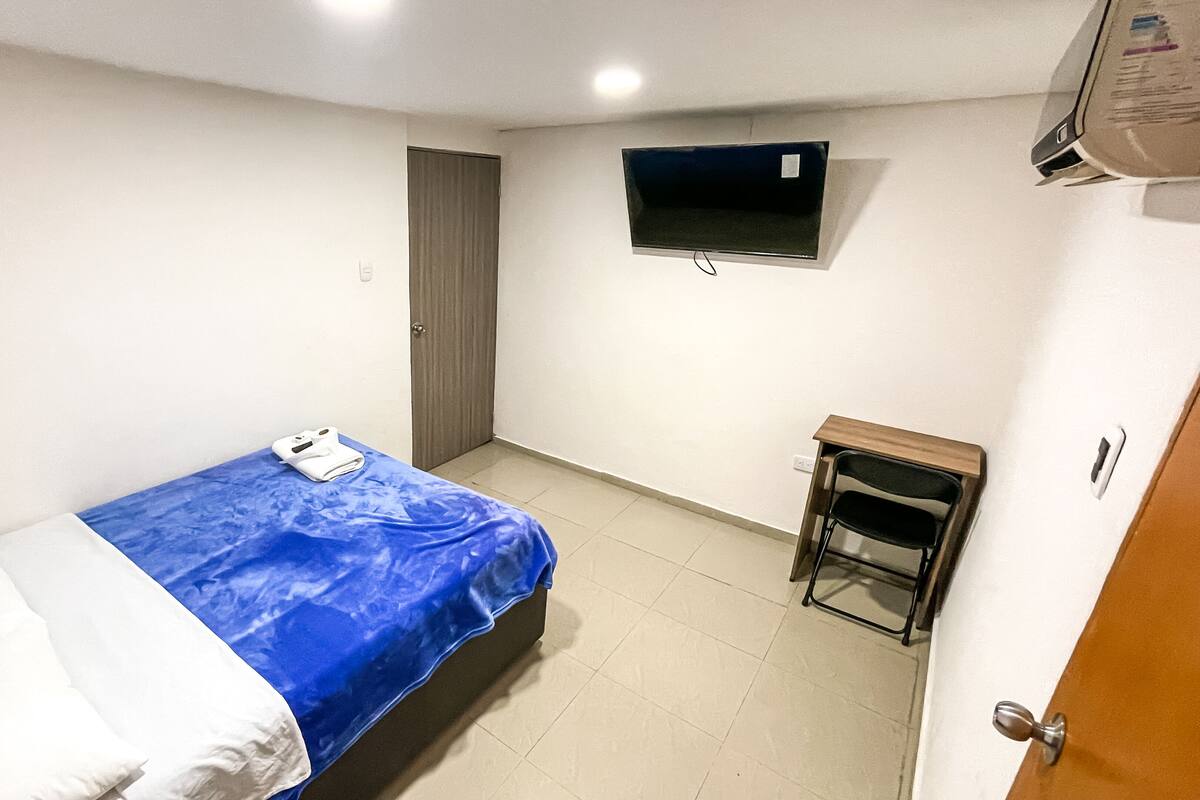 A small, sparsely furnished room with a bed, a television mounted on the wall, and a small table.