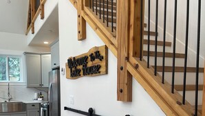 Una cocina moderna con gabinetes grises, un refrigerador de acero inoxidable y una escalera de madera con barandillas negras. Un cartel de bienvenida cuelga en la pared.