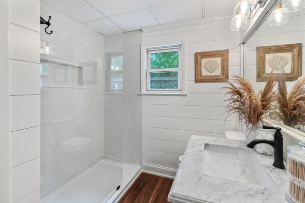 A bathroom with a shower, a vanity with a marble countertop, a window, and decorative shells on the wall.