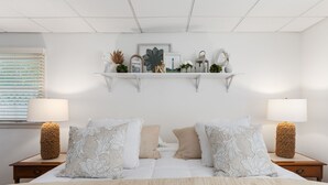 A neatly arranged bedroom with a large bed, two bedside tables with lamps, a wall-mounted shelf with decorative items, and a window with blinds. - Stylish 1BR Apartment in Cape May (Cape May)