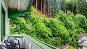 A balcony with chairs, a view of a forested hillside, and a building with a green roof.