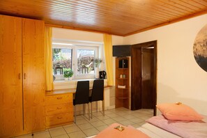 A room with a wooden ceiling, a window with curtains, a desk with two chairs, a bed with pink bedding, and a TV mounted on the wall.