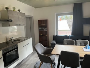 Una cucina moderna con forno integrato, un'area pranzo con tavolo e sedie in legno, una libreria e una finestra con tende.