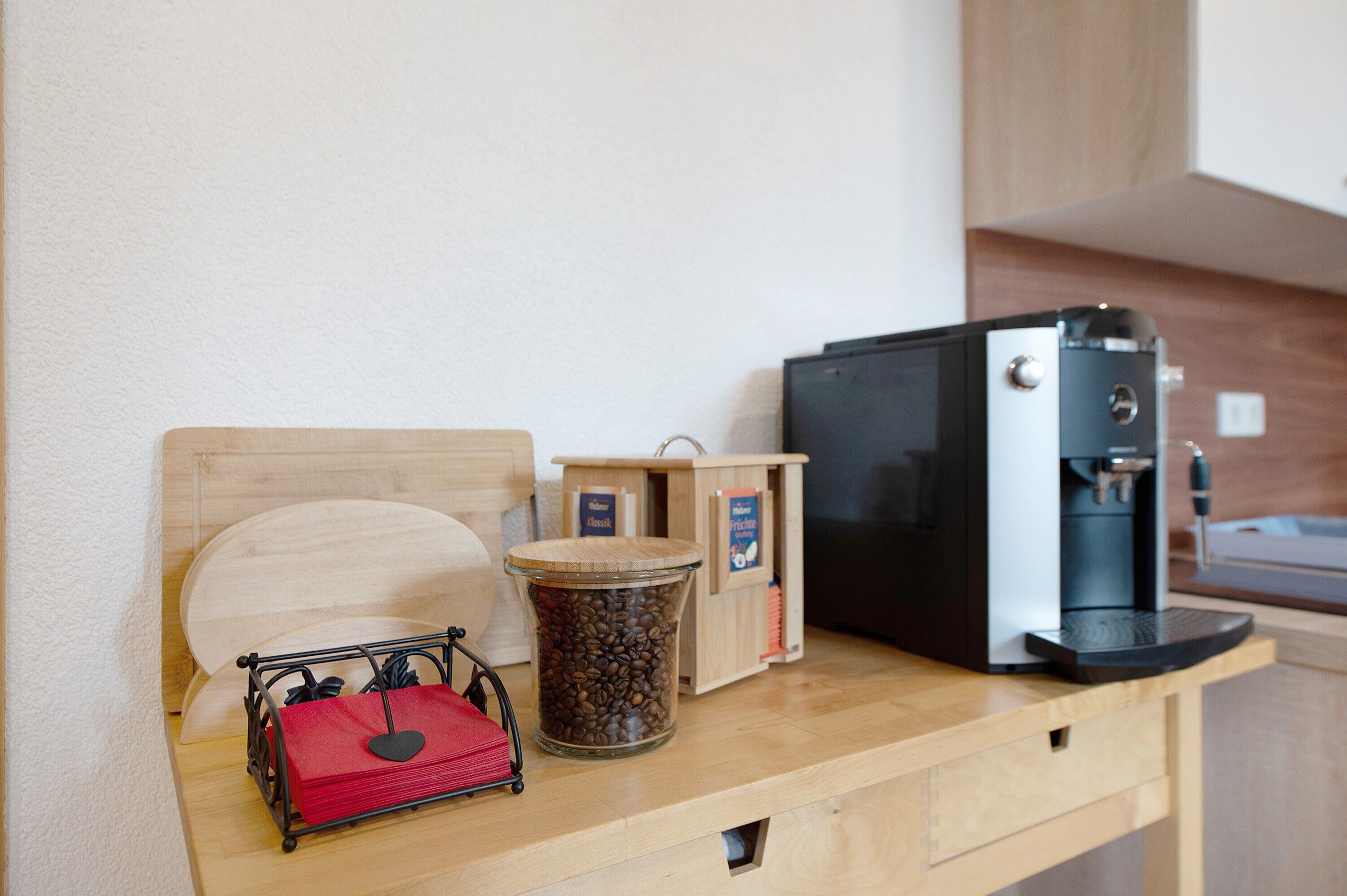 Una kitchenette con cafetera, un recipiente para granos de café, una bolsa roja y un cabecero de madera.