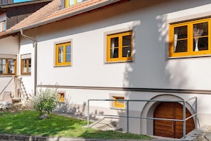 Un bâtiment aux murs blancs et aux fenêtres encadrées de jaune, avec une porte en bois et un petit jardin.