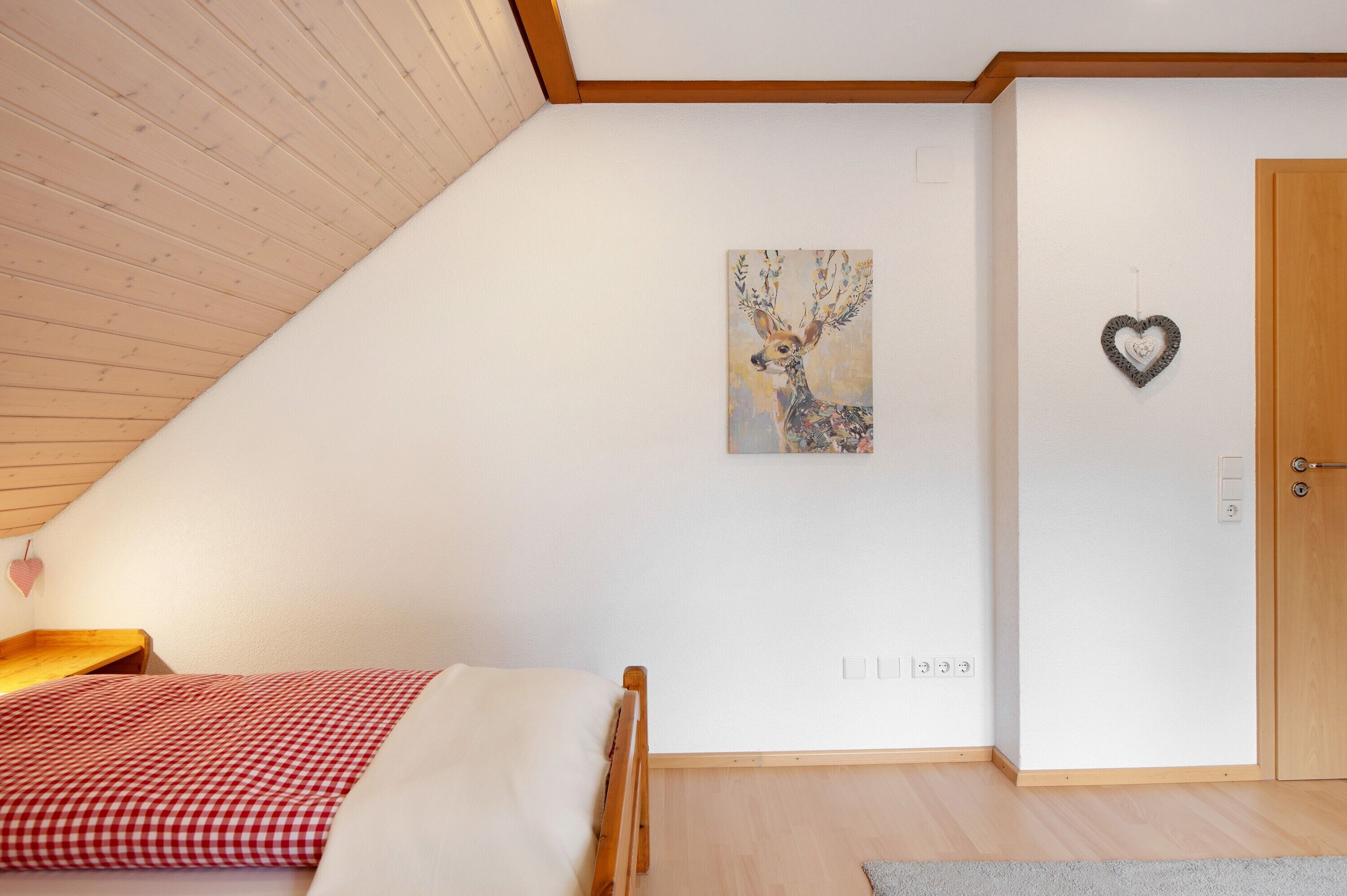 A bedroom with a sloped ceiling, a bed with a checkered bedspread, a painting of a deer, a heart-shaped decoration, and a wooden door.