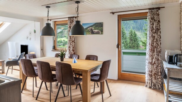 Un comedor con mesa de madera, sillas marrones, lámparas colgantes, un cuadro y vista a un bosque por una ventana.