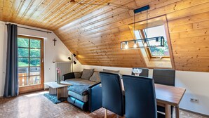Une pièce chaleureuse avec un plafond en pente, un lit, une table à manger avec des chaises et une grande fenêtre donnant sur un paysage verdoyant.
