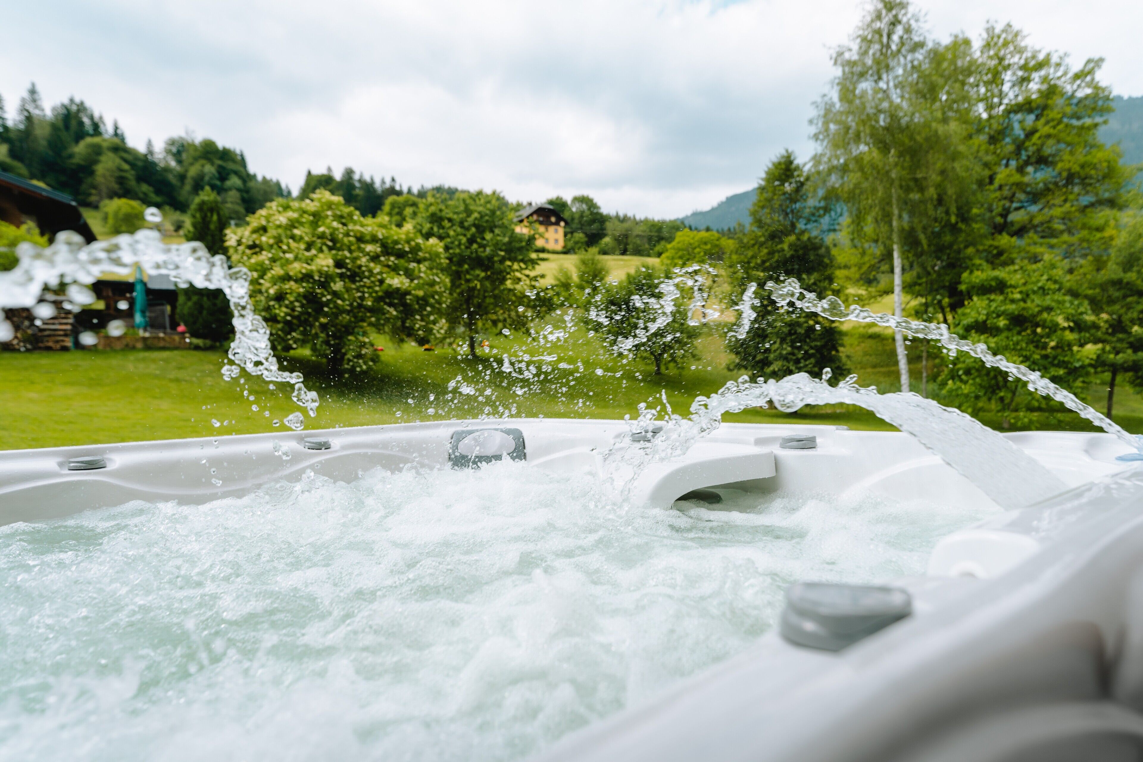 A hot tub with water jets, surrounded by a lush green landscape and a house in the background.