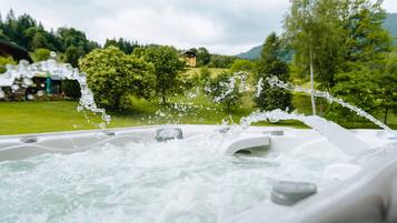 A hot tub with water jets, surrounded by a lush green landscape and a house in the background.
