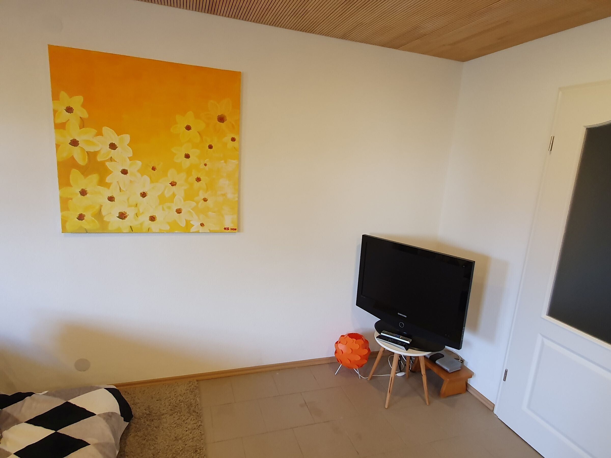 Un salon avec un téléviseur à écran plat posé sur un meuble, un tableau accroché au mur et un petit objet décoratif orange sur le sol.