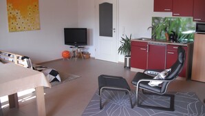 Una sala de estar con televisión, una mesa de comedor, un sofá, una silla y una zona de cocina con armarios y una planta.