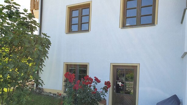Edificio blanco con dos ventanas y una puerta, rodeado de follaje y una silla techada.