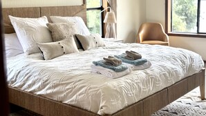 A neatly made bed with a white comforter and several pillows, a wooden headboard, a chair, and a window with curtains.