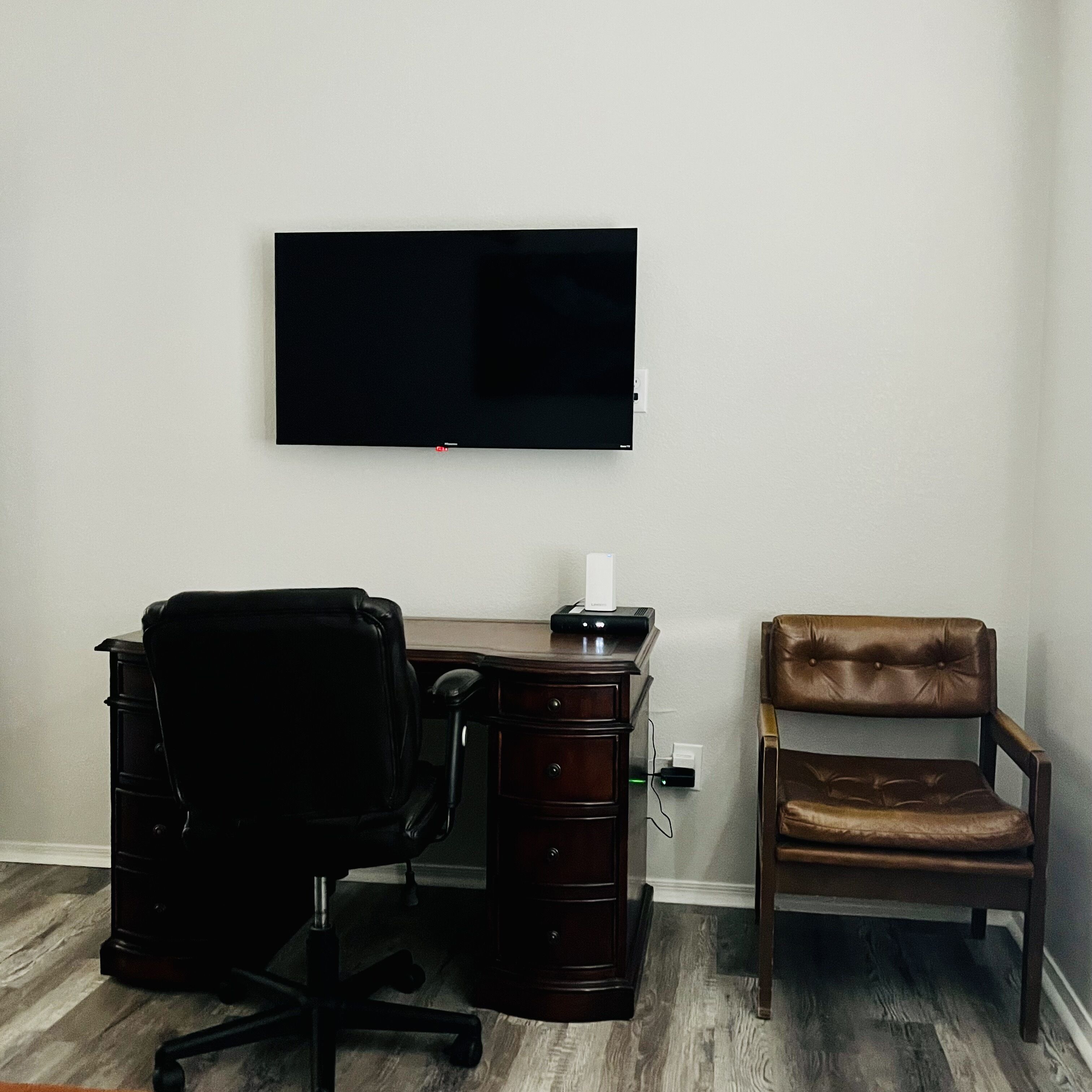 A room with a desk, a chair, a flat-screen TV mounted on the wall, and a power outlet.