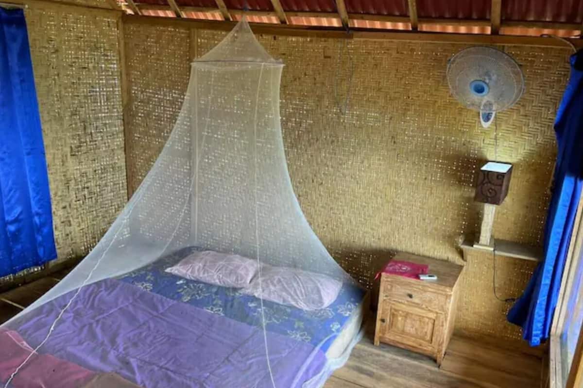 A single bed with a mosquito net, a wooden nightstand, a small table, and a wall fan.