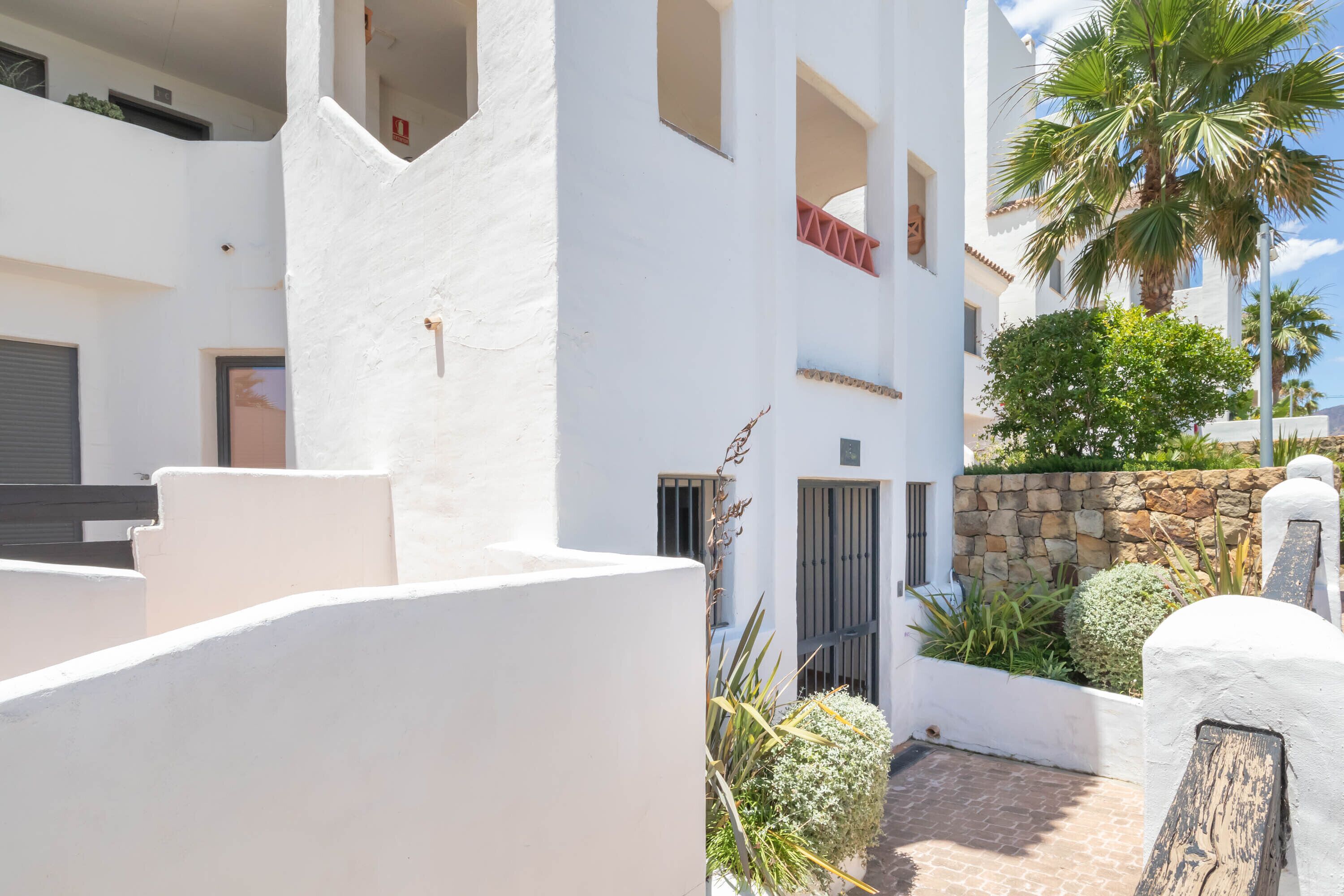 Un edificio blanco con balcones, un muro de piedra y una palmera.