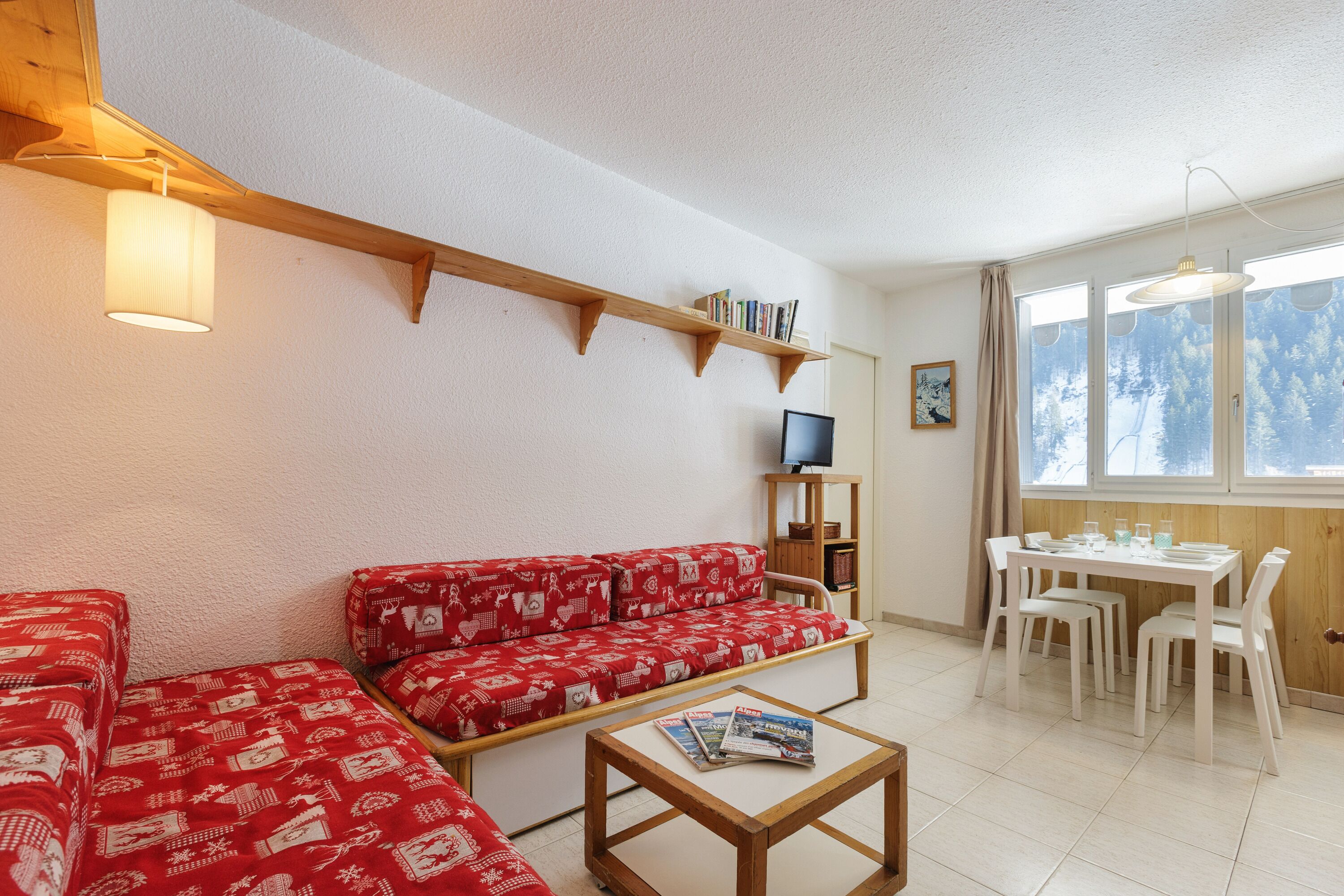 A cozy living room with a red patterned sofa, a wooden coffee table, a TV, and a dining area with a white table and chairs.