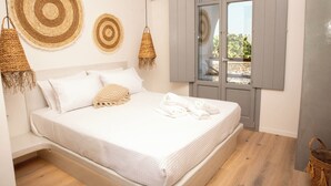 A neatly made bed with white linens, a woven throw blanket, and a pair of towels on the floor. There are two woven wall hangings above the bed and a woven basket hanging from the ceiling. - Aidani Luxurious Rooms  (Santorini)