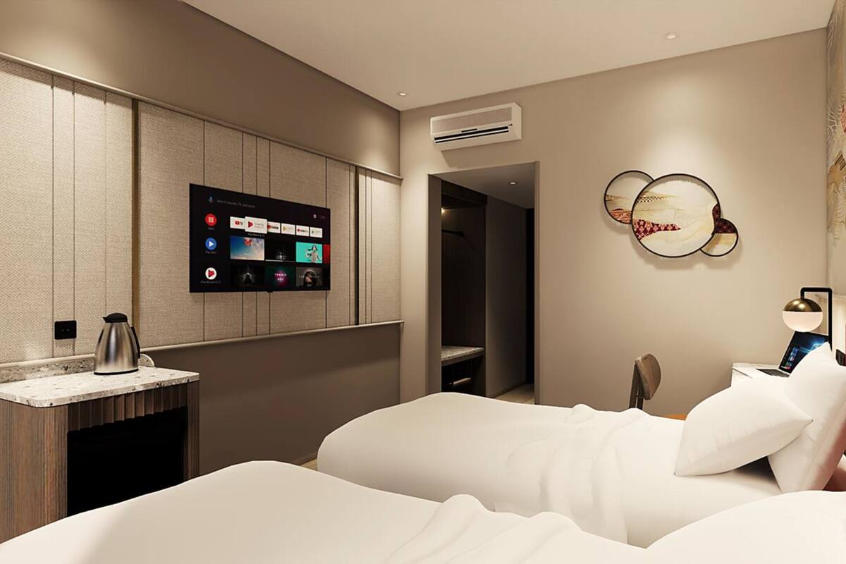 Ein modernes Hotelzimmer mit einem an der Wand befestigten Flachbildfernseher, einem Nachttisch mit Wasserkocher sowie einem Bett mit weißer Bettwäsche und einem dekorativen Wandbild.