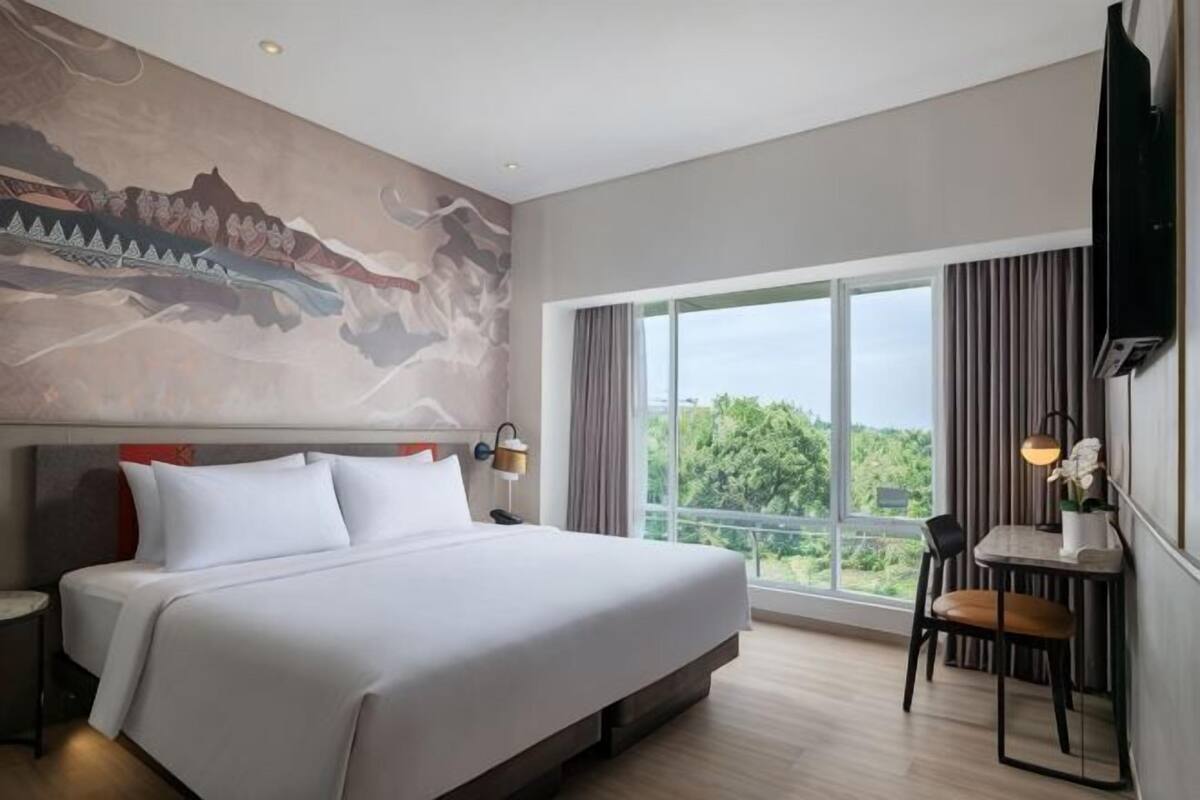 Une chambre d’hôtel dotée d’un grand lit, d’un bureau avec une chaise, d’un téléviseur fixé au mur et d’une grande fenêtre donnant sur un paysage verdoyant.