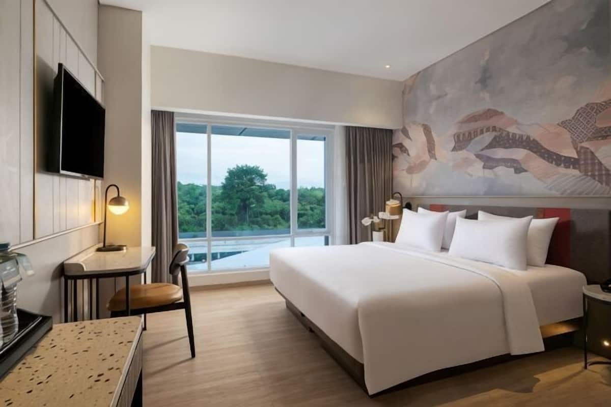 Ein Hotelzimmer mit einem großen Bett, einem Schreibtisch, einem Sessel, einem Fernseher und einem großen Fenster mit Blick auf Bäume.