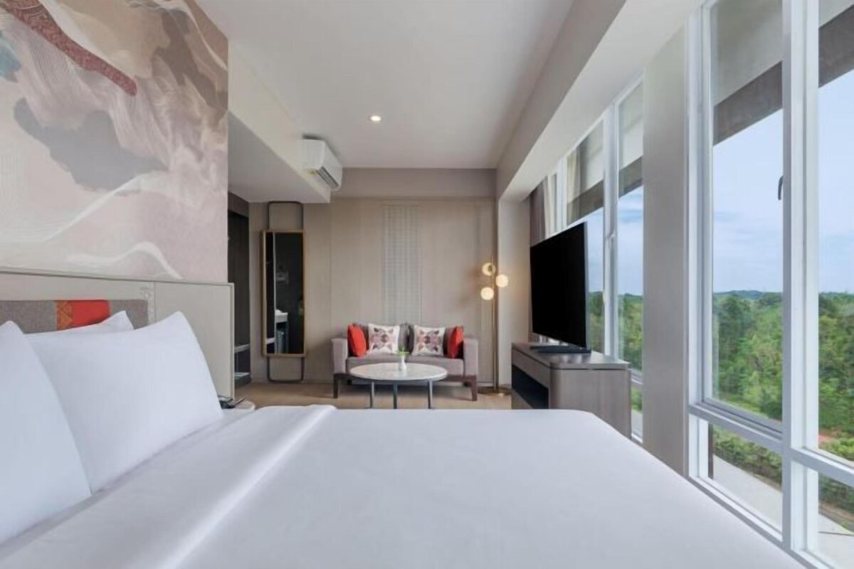 A modern hotel room with a large bed, a seating area with a table and chairs, a flat-screen TV, and a large window offering a view of the outdoors.