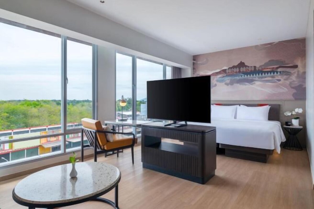 A modern hotel room with a large window, a flat-screen TV, a bed, a small table, and a chair.