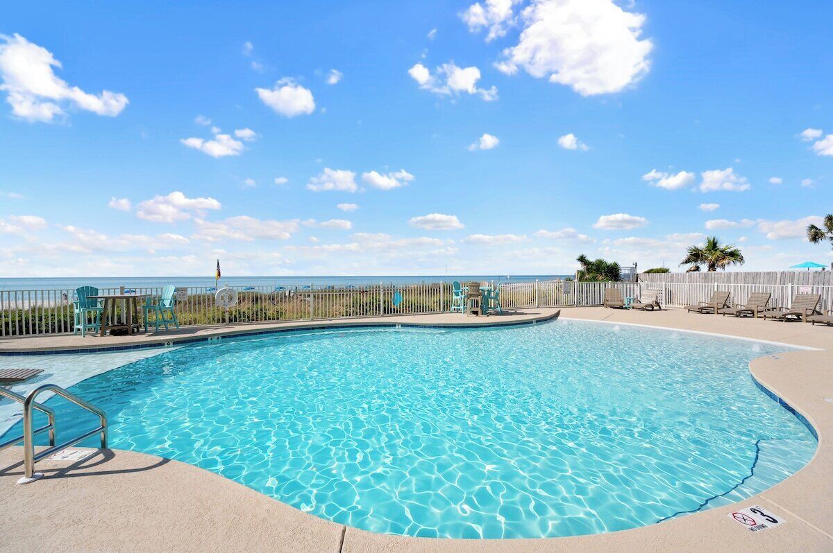 A clear blue pool with a wooden deck, lounge chairs, and a view of the ocean.