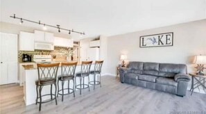 Una cocina moderna con armarios blancos, una zona de bar con taburetes y un sofá de cuero gris en la sala de estar.
