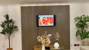 Un salon moderne avec un téléviseur à écran plat fixé sur un mur recouvert de panneaux en bois, une table basse ronde en verre, une plante en pot et un canapé.