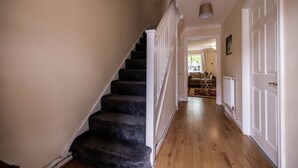 Un escalier dont les marches sont recouvertes de moquette mène à un palier. Une porte blanche est visible sur la droite, et un couloir s’étend vers le fond.
