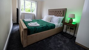 Un lit bien fait, avec une couvre-lit verte et des oreillers blancs, une lampe de chevet et une fenêtre avec des rideaux.