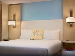 Quarto de hotel com cama, duas luminárias de cabeceira, mesa de cabeceira com um tablet e um livro, e parede em tons de azul e bege.