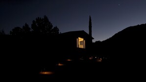 A small, dark house with a lit window, surrounded by trees and a hill.