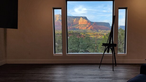 A room with a large window overlooking a mountainous landscape, a wooden floor, and a black easel.