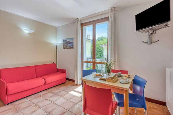 Une pièce avec un canapé rouge, une table à manger en bois entourée de chaises bleues, un téléviseur fixé au mur et une fenêtre avec des rideaux.
