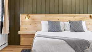 Ein ordentlich bezogenes Bett mit weißer Bettwäsche und gemusterten Kissen, ein Holz-Kopfende und ein Nachttisch mit Lampe.