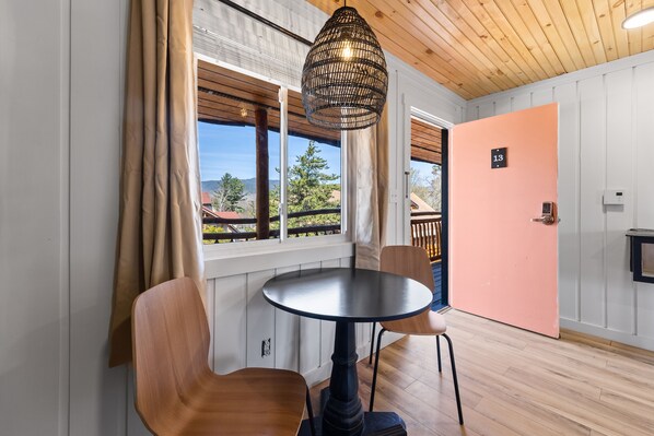 A room with a round table, two chairs, a hanging light fixture, a door with a number, and a window with curtains.