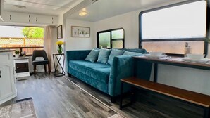 Interior de una caravana compacta con kitchenette, un sofá azul, una mesa de comedor y una ventana con vistas.