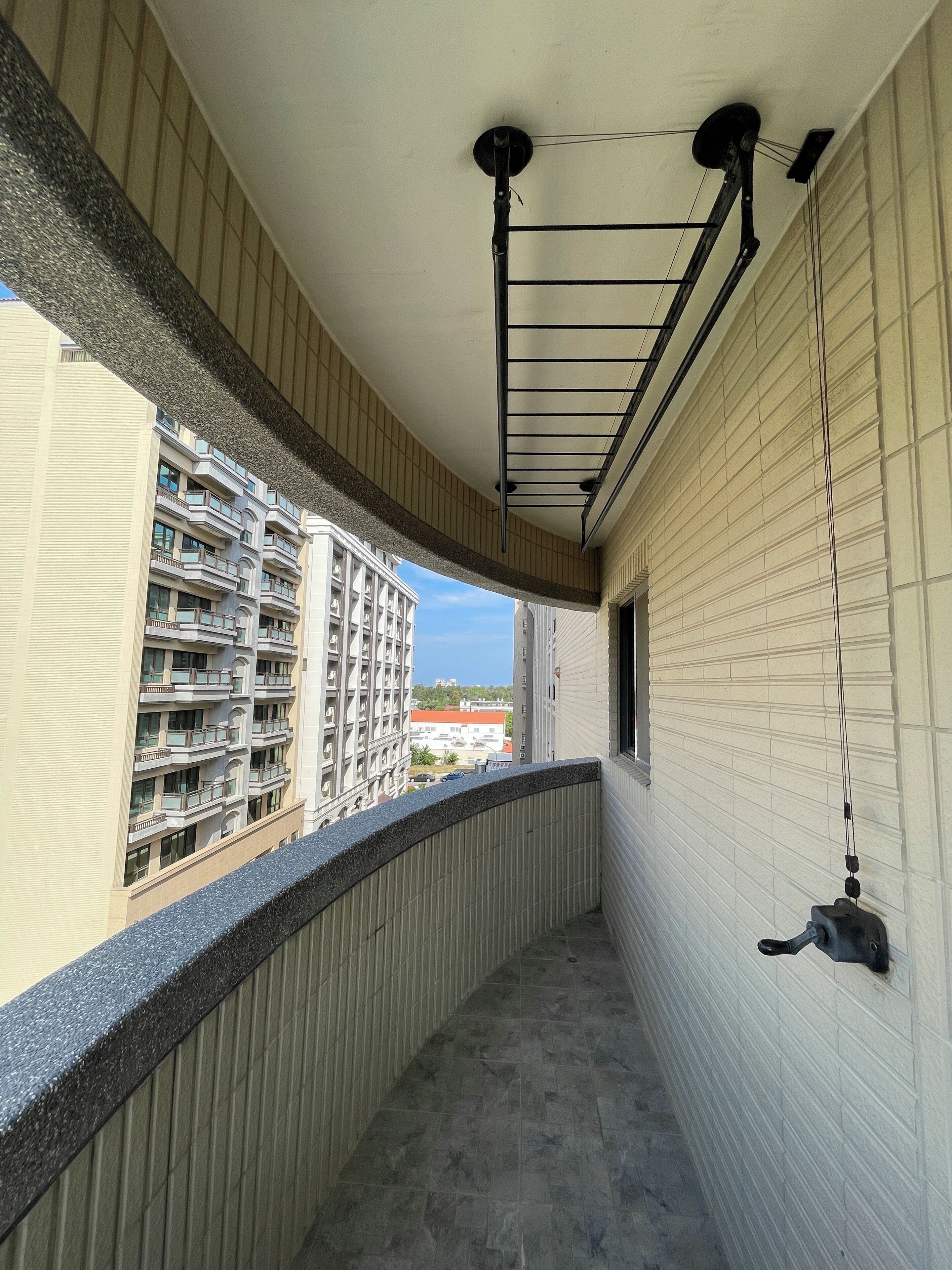 Un estrecho pasillo con suelo de baldosas, una barandilla metálica en un lado y vista de un edificio de varios pisos con balcones.