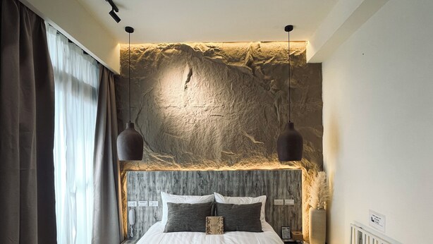 Une chambre moderne avec un grand lit, deux lampes suspendues, un mur à texture et une vue sur l’extérieur à travers des rideaux légers.