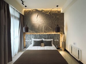 Un dormitorio moderno con una cama grande, dos lámparas colgantes, una pared con textura y vistas al exterior a través de cortinas traslúcidas.
