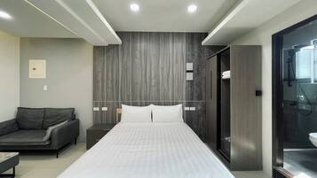 Una habitación de hotel moderna con una cama grande, un sofá oscuro, cabina de ducha de vidrio y un diseño minimalista.