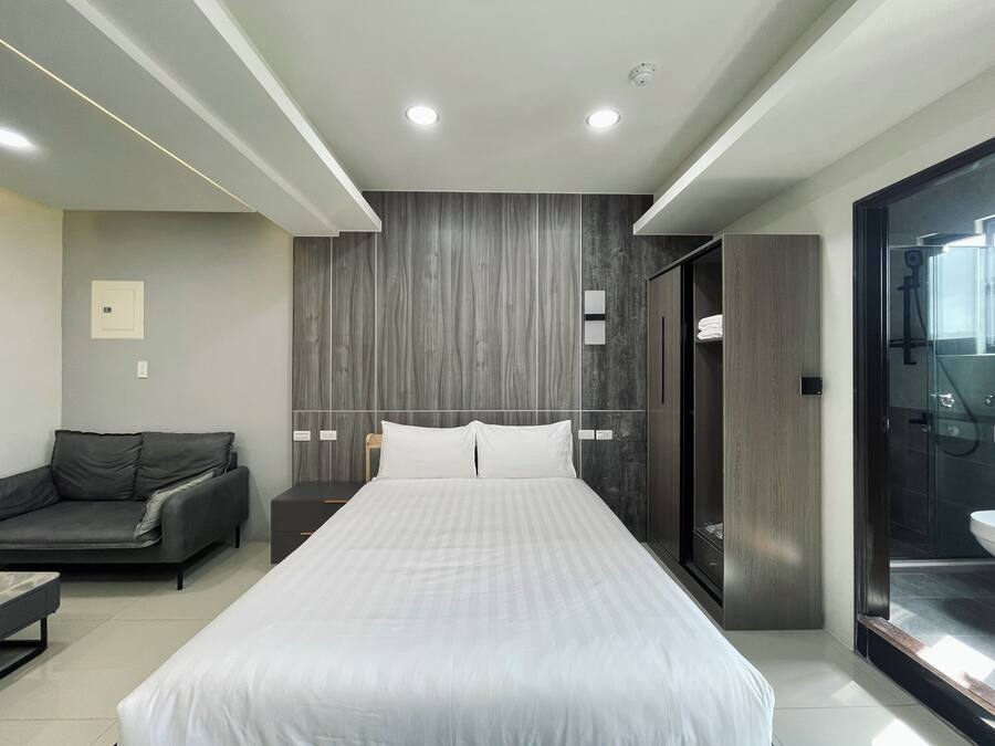 Una habitación de hotel moderna con una cama grande, un sofá oscuro, cabina de ducha de vidrio y un diseño minimalista.